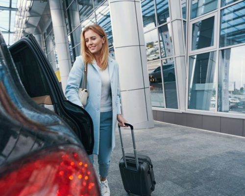 Smiling pleased woman with trolley suitcase closing door of taxicab parked before airport terminal building